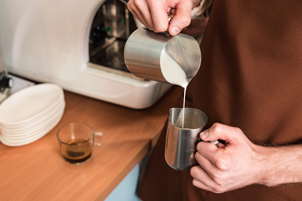 barista with brown apron pouring milk to a metal jug