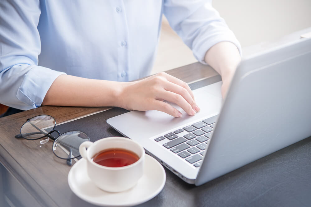 woman in blue shirt typing something in her laptop with a cup of tea and sunglasses besides her