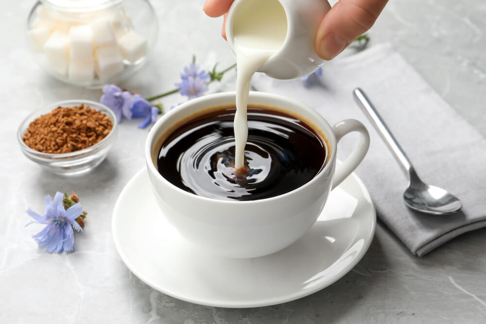 Woman pouring milk into a cup with a delicious chicory drink at a light grey marble table