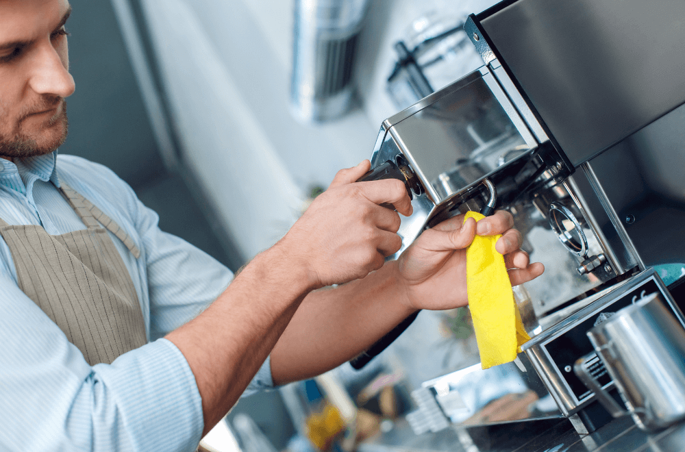 A young handsome man barista standing at a coffee shop cleaning a steamer with a wipe concentrated close-up