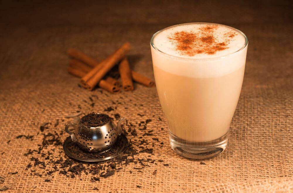 A glass of dirty chai latte with chai tea leaves and cinnamon beside