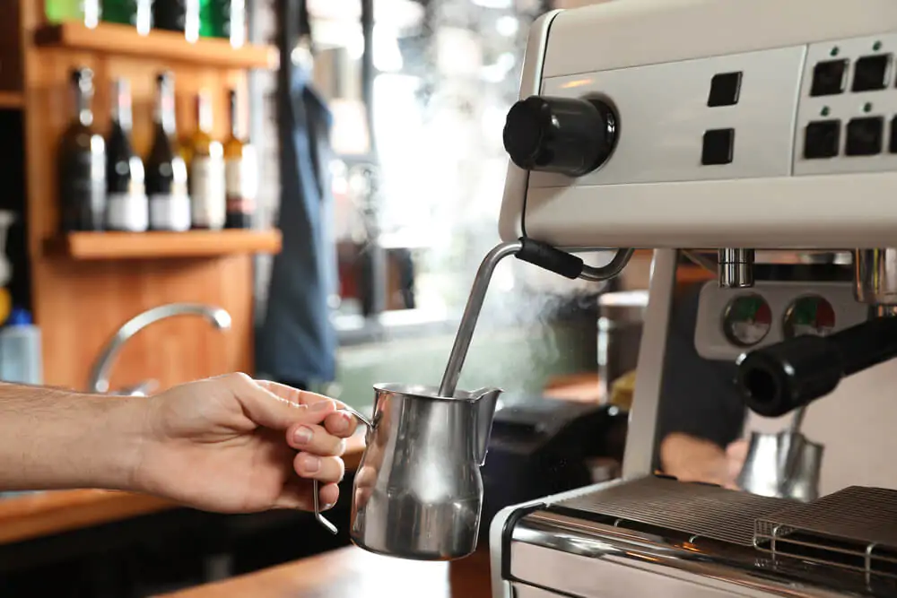 Barista steaming milk in metal jug with coffee machine wand at bar counter