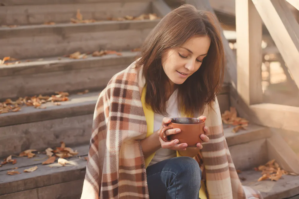 A young woman sitting on the stairs with autumn leaves drinking a hot beverage.