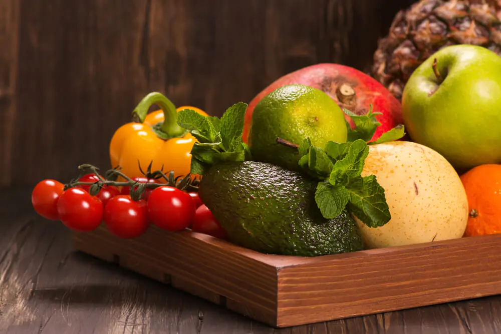 A wooden container containing green apple, avocado, red tomatoes, orange bell pepper.