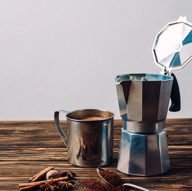 an open moka pot and a mug of coffee beside it with raw ingredients on a wooden table