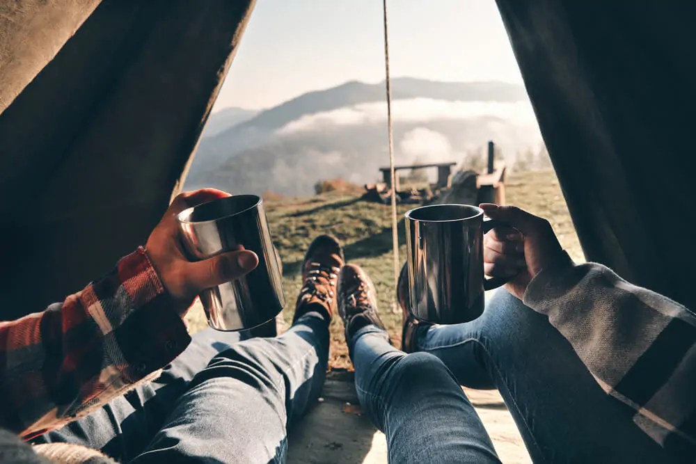 close up of couple enjoying view of mountain range while lying in tent with hot drinks in mugs