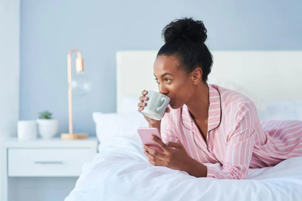 Young woman drinking coffee while using her cellphone in bed at home.