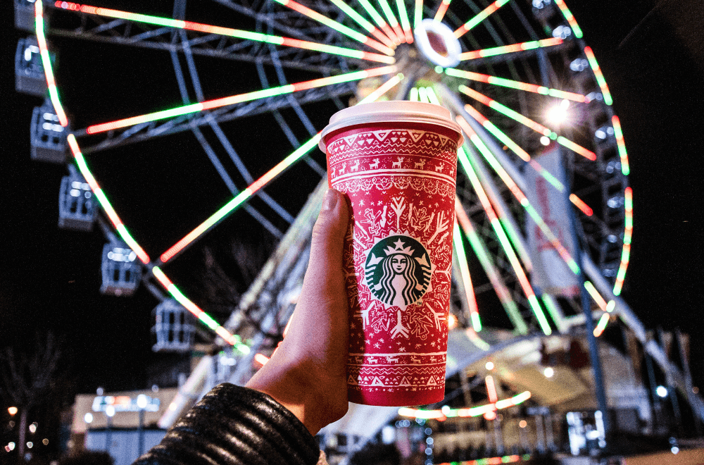 A man holding his Starbucks holiday cups up with a Ferris wheel at the background