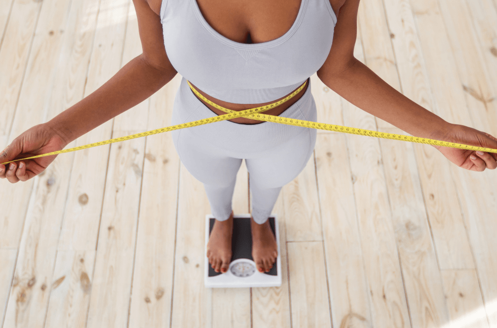 Above view of African American lady measuring her waist with tape, standing on scales indoors, closeup