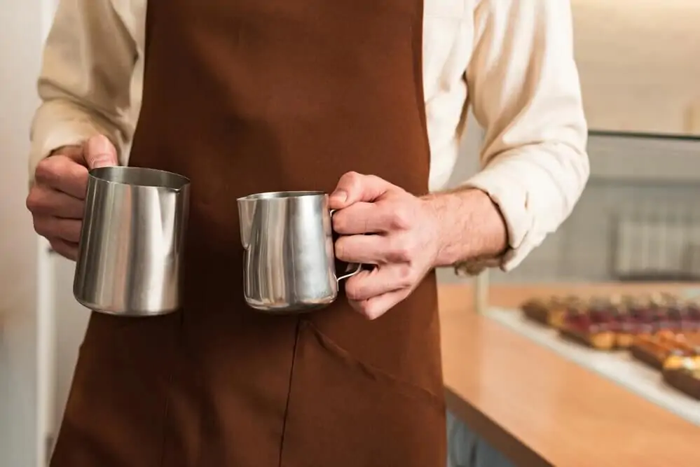 barista in a brown apron holding steel milk jugs