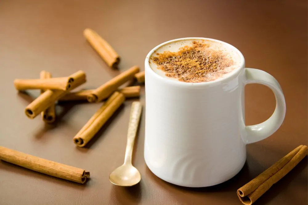 A plain white cup of hot coffee topped with cinnamon powder, decorated with cinnamon sticks