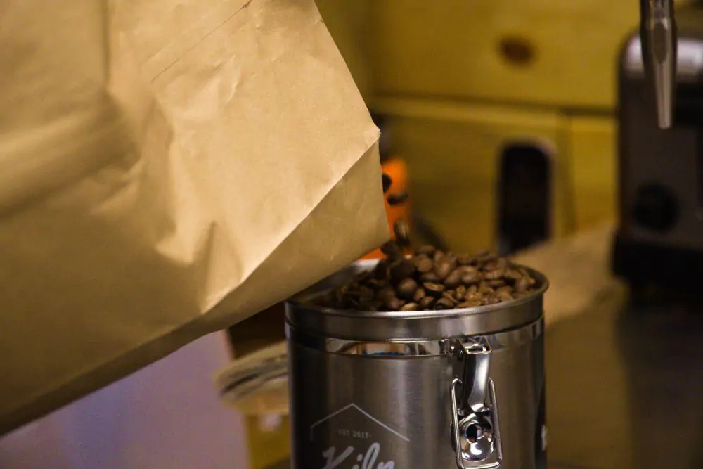 A metal canister is filled with raw coffee beans from brown paper bag