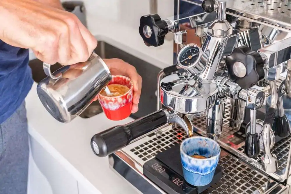 Barista man making cortado coffee at coffee machine pouring milk in espresso cup at cafe