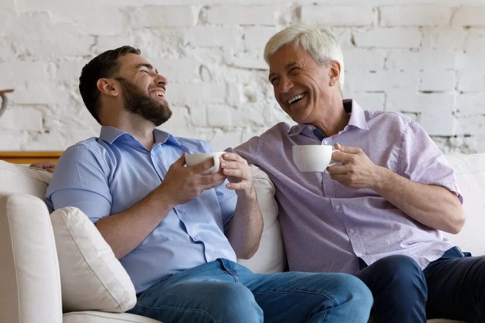 Happy adult son meeting with mature retired grey haired father, drinking hot tea, coffee, talking, laughing, enjoying family leisure time on weekend at home, keeping good healthy warm relationships