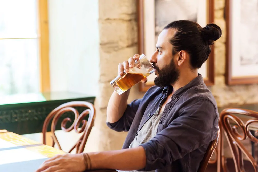 people, drinks, alcohol and leisure concept - happy young man drinking beer from glass at bar or pub