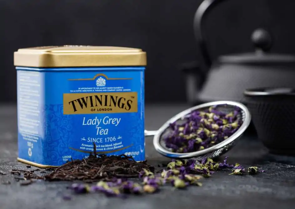 Steel jar of Twinings Lady Grey loose tea with iron teapot and blue mallow flowers on black background
