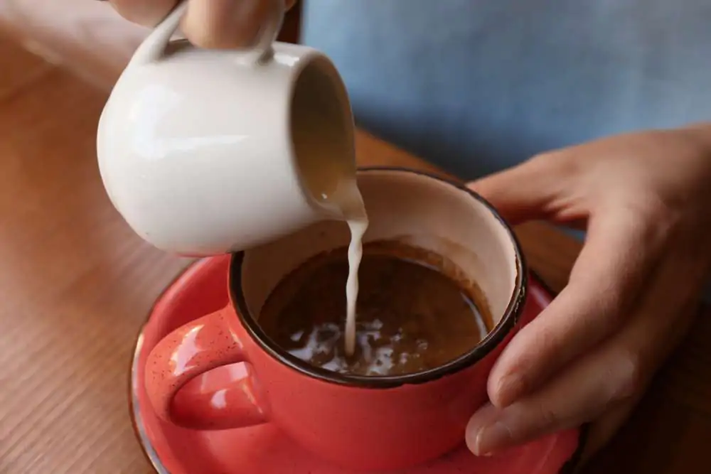Woman adding milk to fresh aromatic coffee at table, closeup